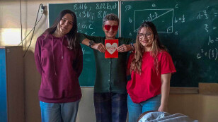 Trzy kobiety na tle zielonej szkolnej tablicy, na tablicy wzory matematyczne, środkowa kobieta ma okulary w kształcie serc, w ręku trzyma serce z napisem